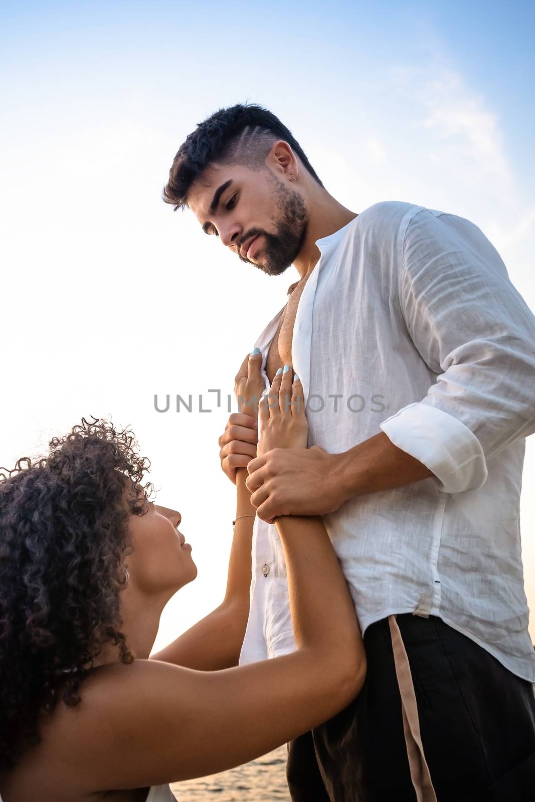 Kneeling submissive woman begs her man by holding his hands on his massive chest, but he squeezes her wrists as a sign of possession and domination - Multiracial lovers couple romance scene by robbyfontanesi