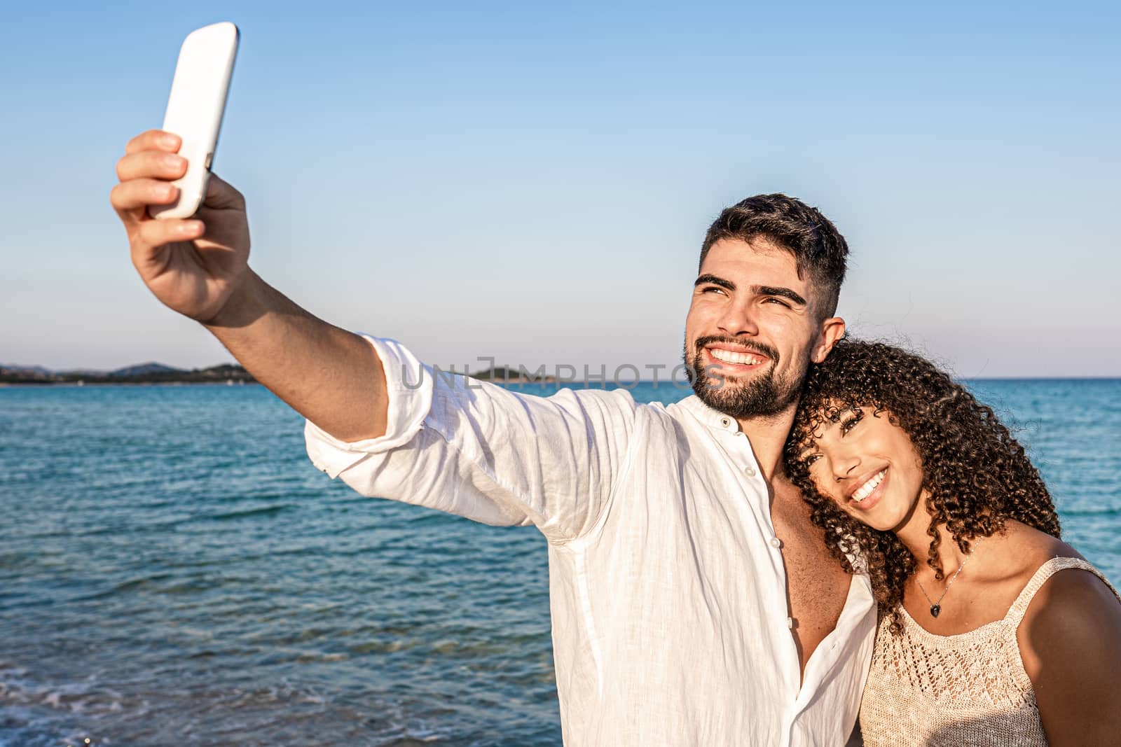 Young mixed race lovers couple at the sunset standing in the seashore makes a self portrait with the smartphone - Caucasian man having fun shooting photo in vacation with her black Hispanic girlfriend