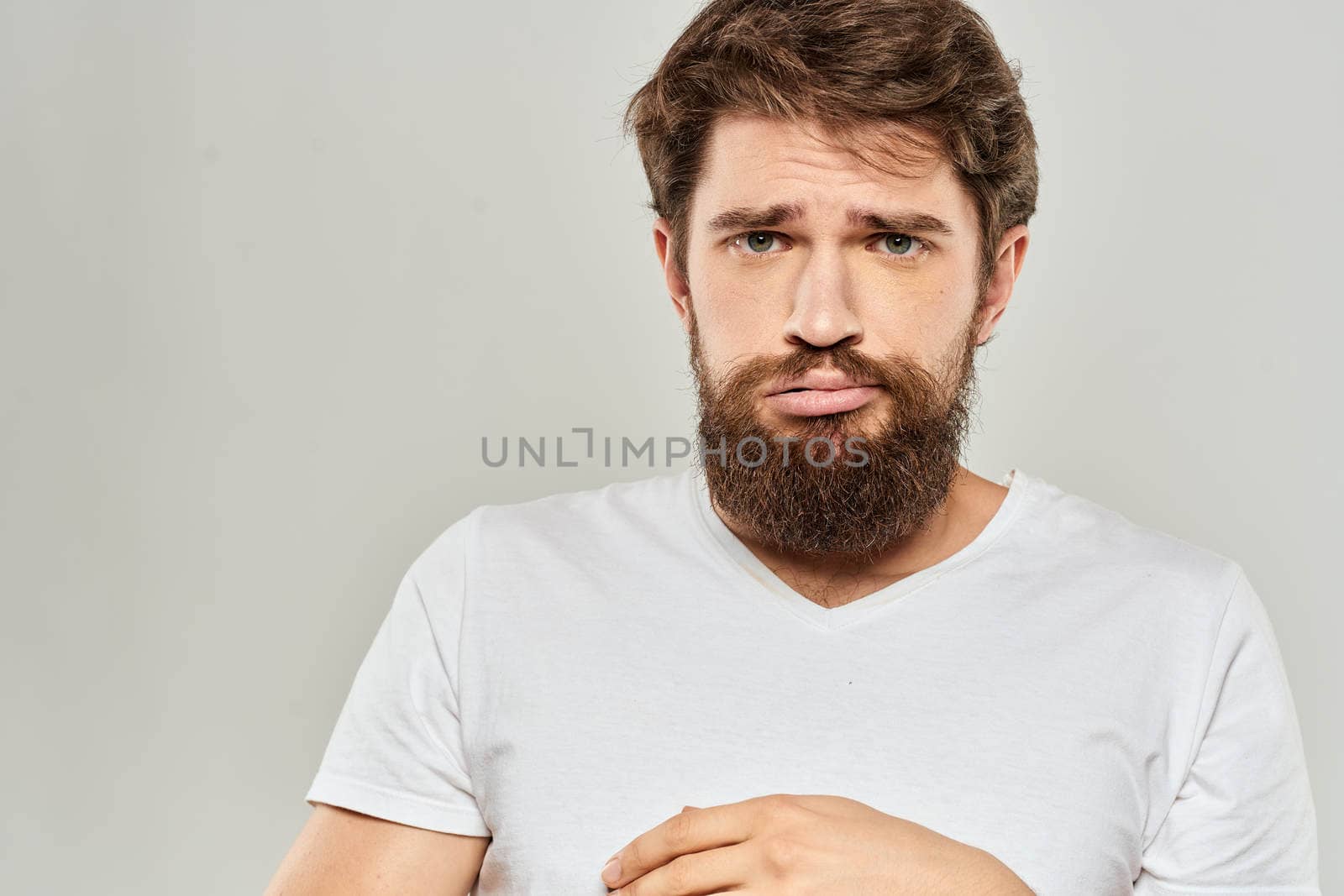 A man in a white t-shirt with a beard emotions displeased facial expression light background. High quality photo