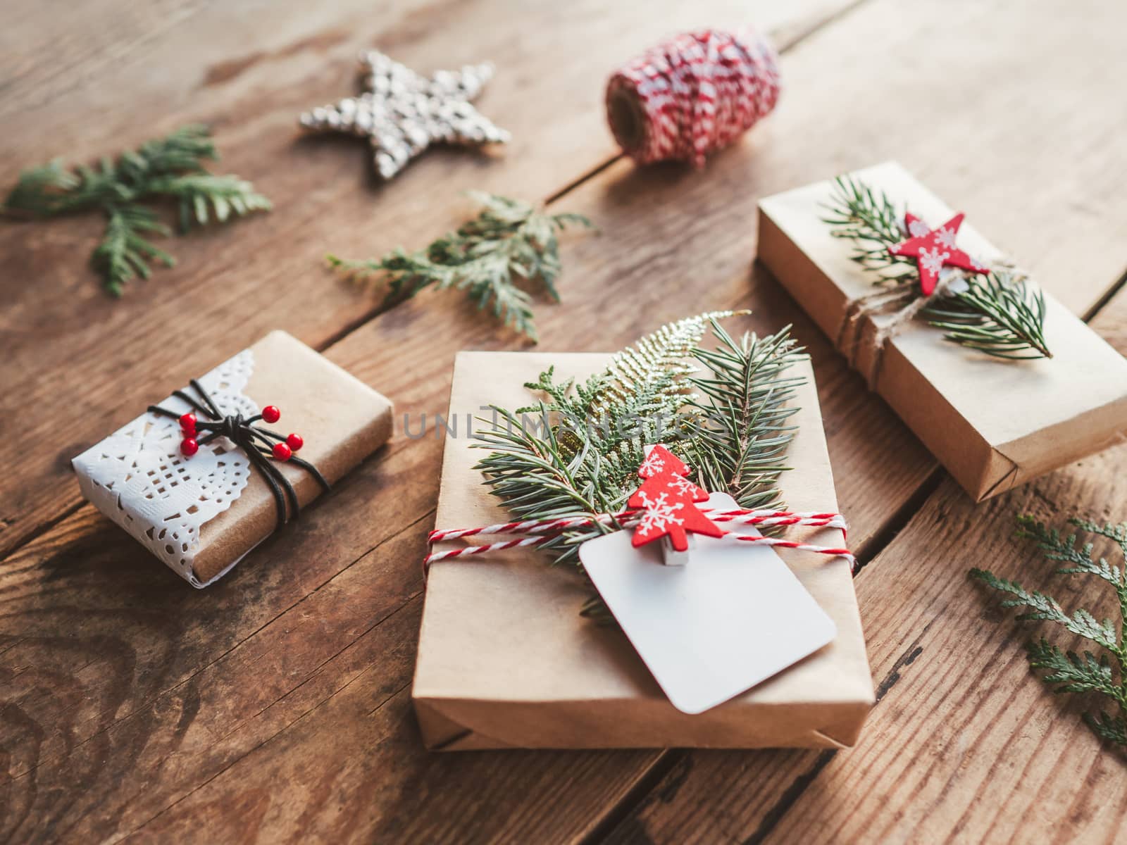 Christmas DIY presents wrapped in craft paper with fir tree twigs. Red decorations in shape of Christmas tree, stars. New Year gifts on wooden background. Winter holiday spirit.