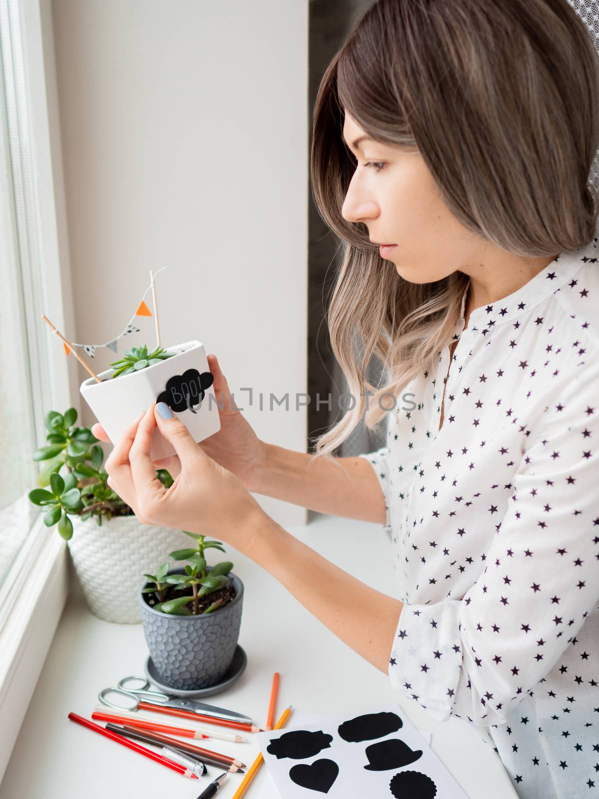 Smiling woman shows handmade decorations for Halloween. DIY flags and Boo! sticker on flowerpot with succulent plant. Socially-Distanced Halloween at home.