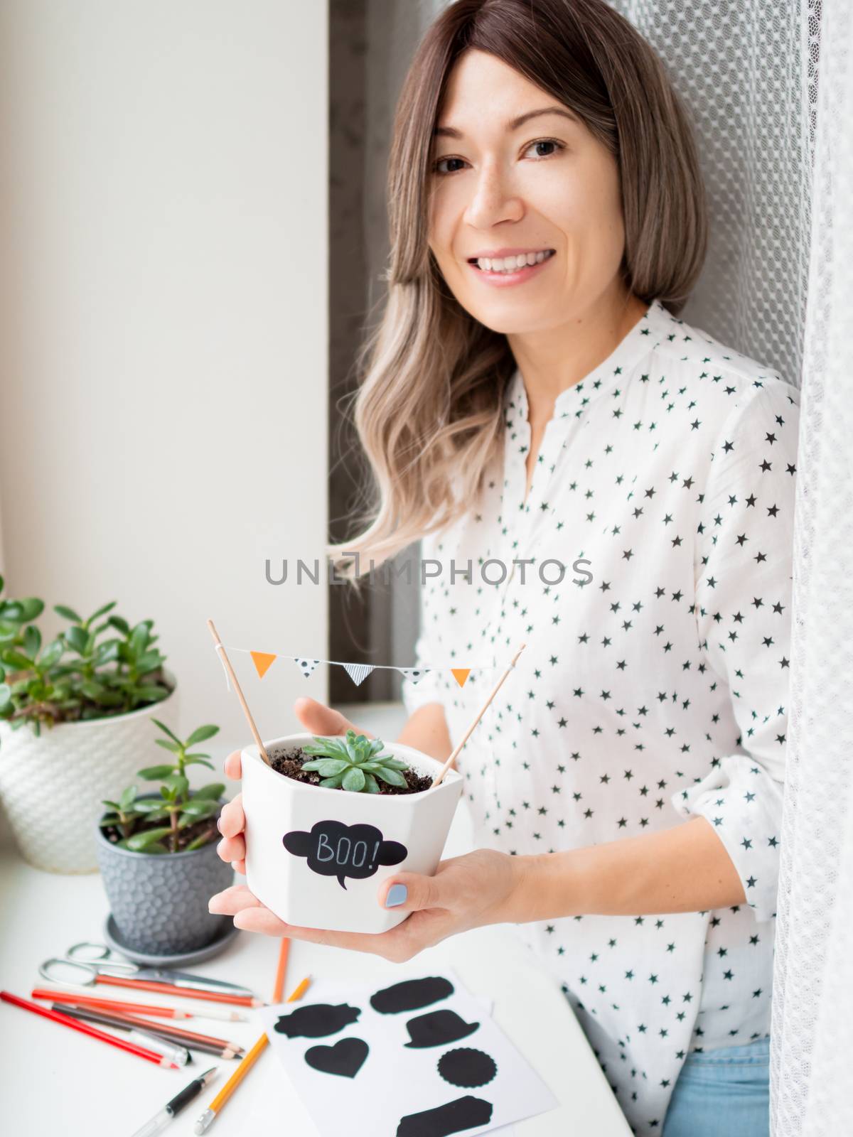 Smiling woman shows handmade decorations for Halloween. DIY flags and Boo! sticker on flowerpot with succulent plant. Socially-Distanced Halloween at home.