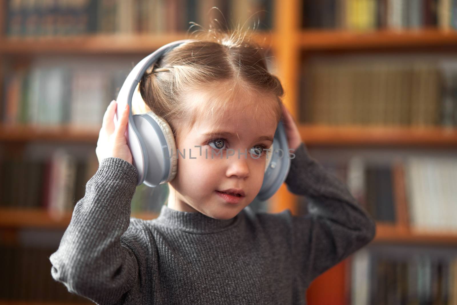 Cute Caucasian little girl in big white headphones listens music in the interior against the background of bookshelves Modern children. Happy childhood with technology Child spends free time at home.