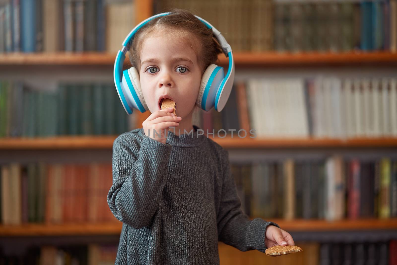 Cute Caucasian little girl in big white headphones listens music in the interior against the background of bookshelves Modern children. Happy childhood with technology Child spends free time at home.