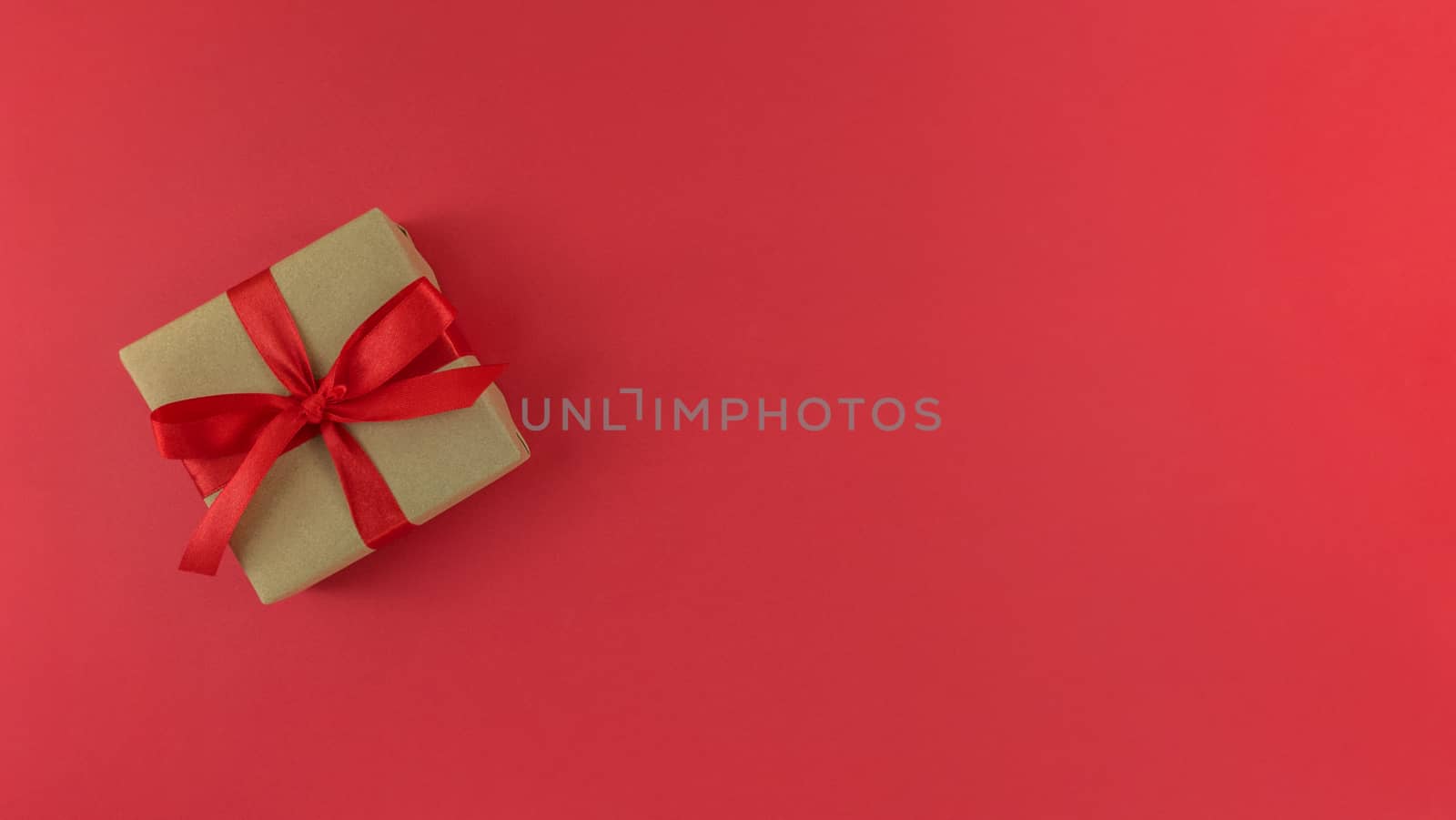 Gift box wrapped in craft paper with red ribbon and bow on red background. Monochrome festive flat lay with copy space.