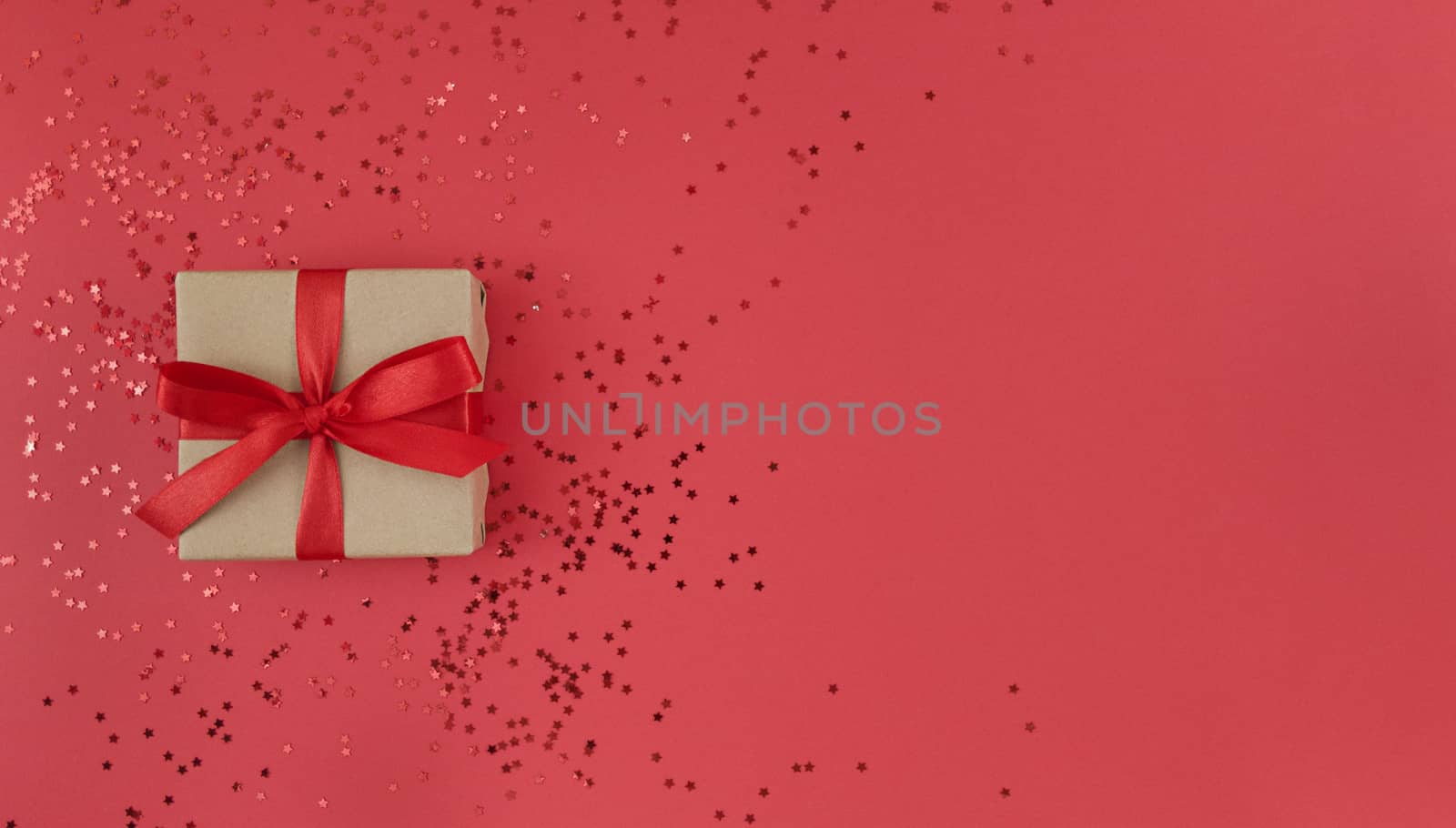 Gift box wrapped in craft paper with red ribbon with bow and confetti on red background. Monochrome festive flat lay with copy space.