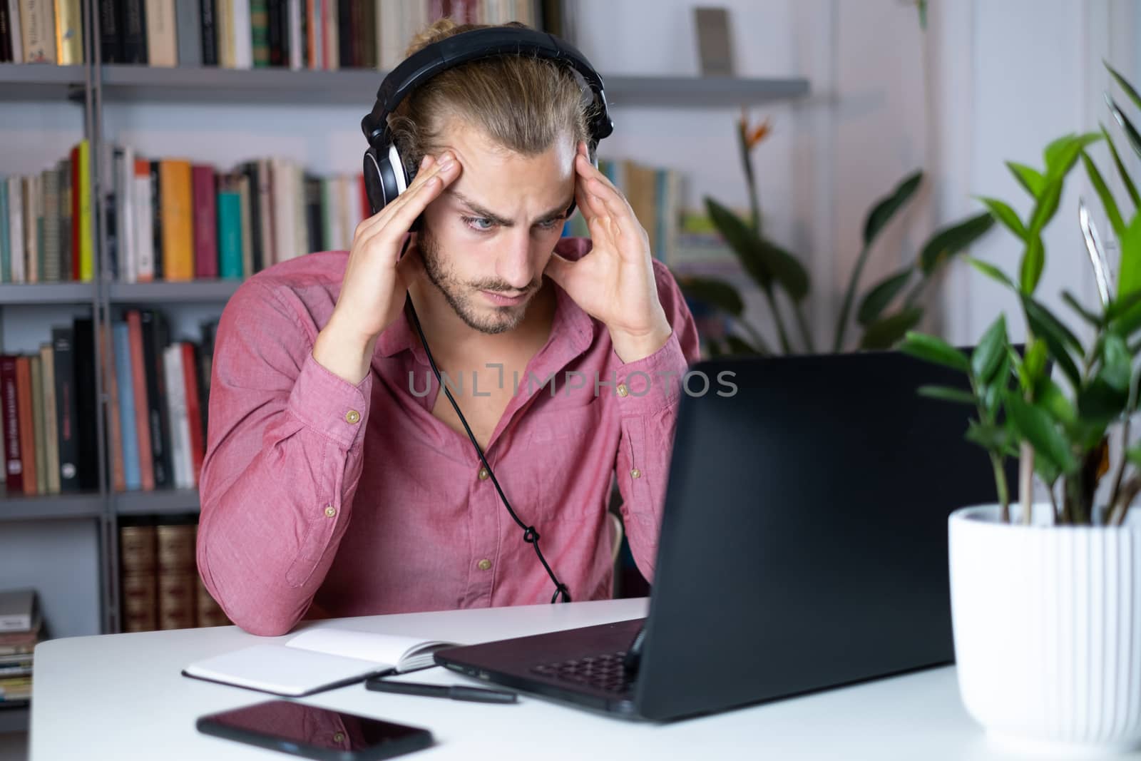 Handsome young caucasian man using computer working at home and talking with headphones feeling worried with hands on head