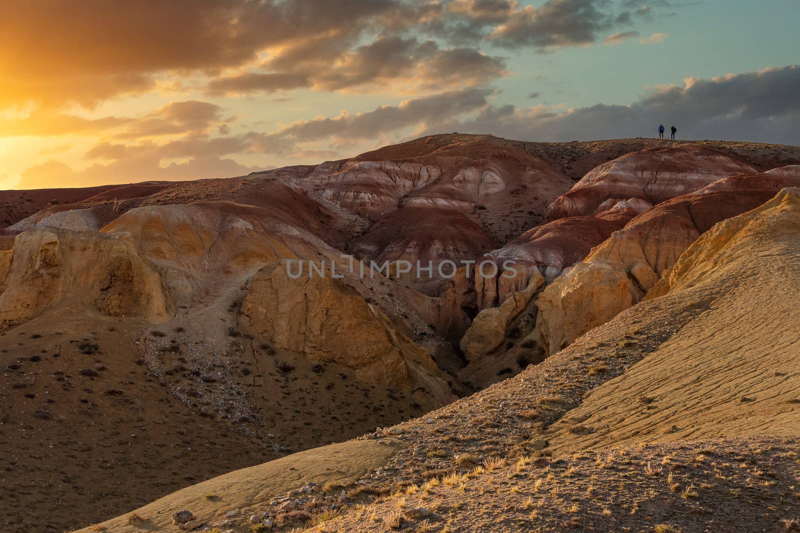 Beautiful low angle shot of two tiny human figures climbing massive red mountain in Kyzyl-Chin valley, also called Mars valley. Sunset cloudy sky as a backdrop. Golden hour. Altai, Siberia, Russia by DamantisZ