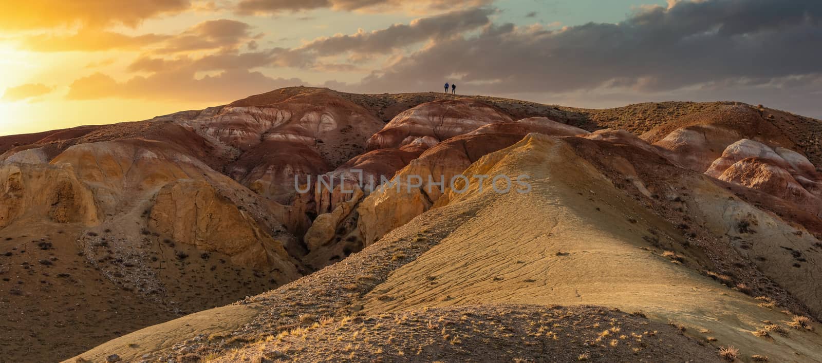 Low angle panoramic shot of two tiny figures on top of massive red mountain in Kyzyl-Chin valley, also called Mars valley. Beautiful sunset sky as a background. Golden hour. Altai, Siberia, Russia by DamantisZ