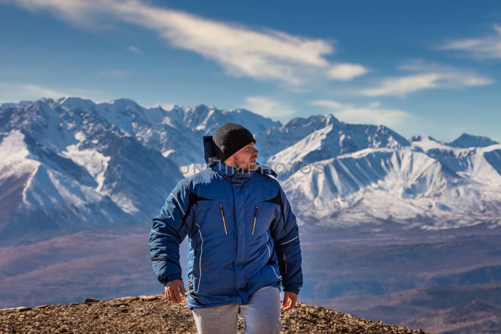 View of a tourist walking in the mountains. White snowy mountain ridge and beautiful blue cloudy sky as a background and slightly out of focus. Altai mountains, Siberia, Russia.