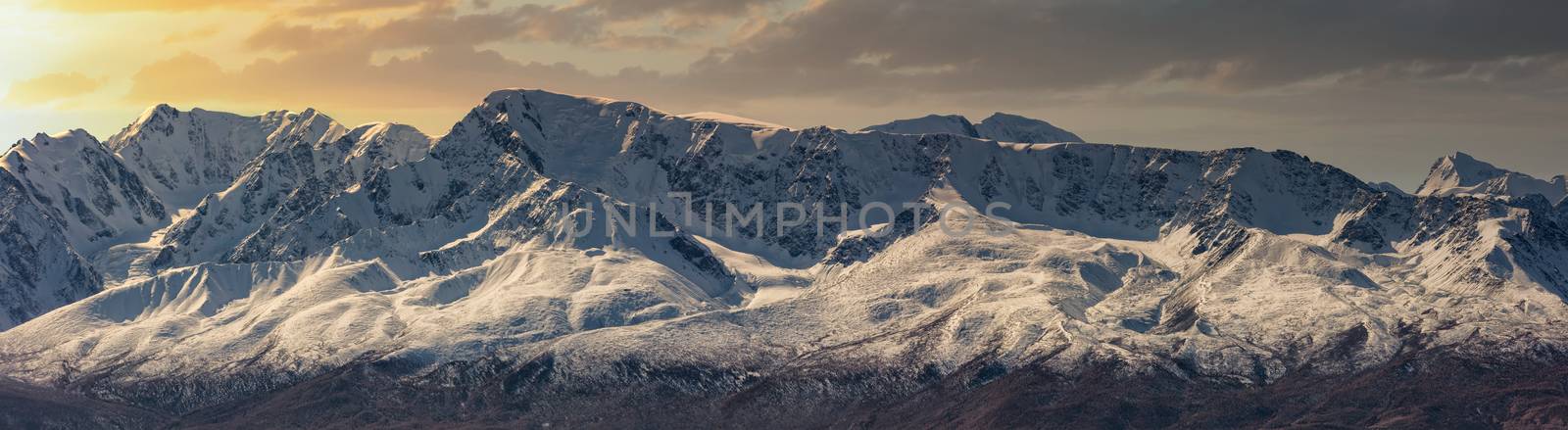 Scenic panoramic aerial view of snowy mountain peaks and slopes of North Chuyskiy ridge at sunset. Beautiful cloudy orange sky as a background. Altai mountains, Siberia, Russia.