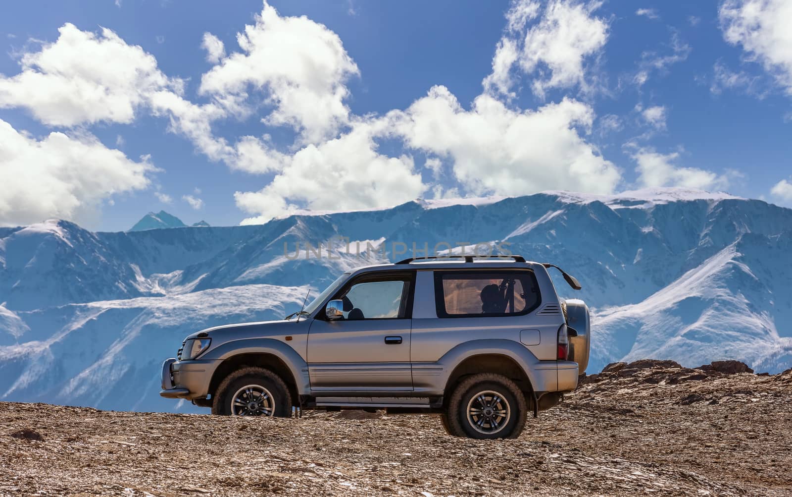 Scenic view of an off-road vehicle parked in the mountains. White snowy mountain ridge and beautiful blue sky as a background slightly out of focus. Altai mountains, Siberia, Russia.