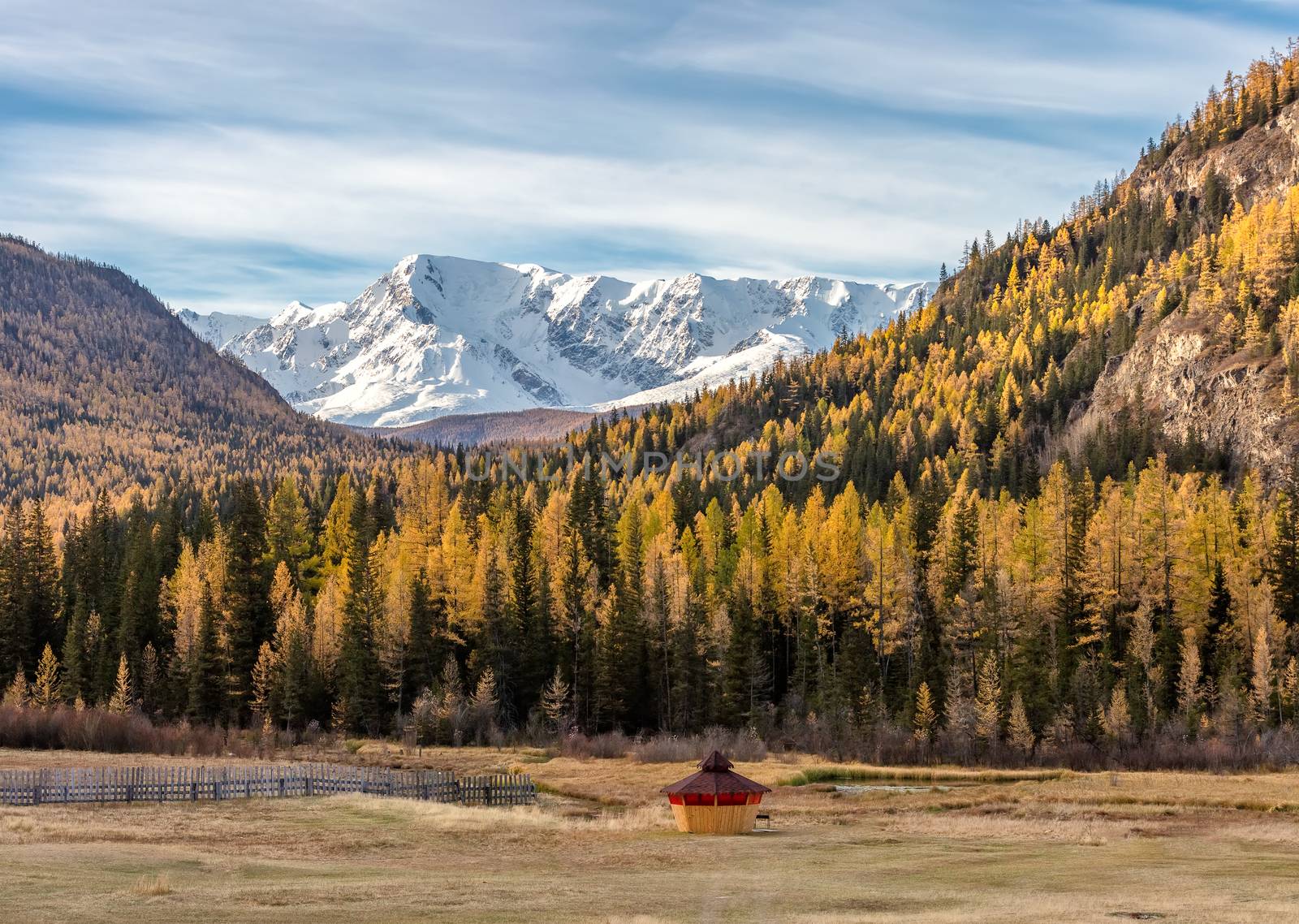 Scenic low angle view of snowy mountain peaks and slopes of North Chuyskiy ridge. Golden trees, wooden hut in the foreground. Beautiful blue cloudy sky as a backdrop. Altai mountains, Siberia, Russia by DamantisZ