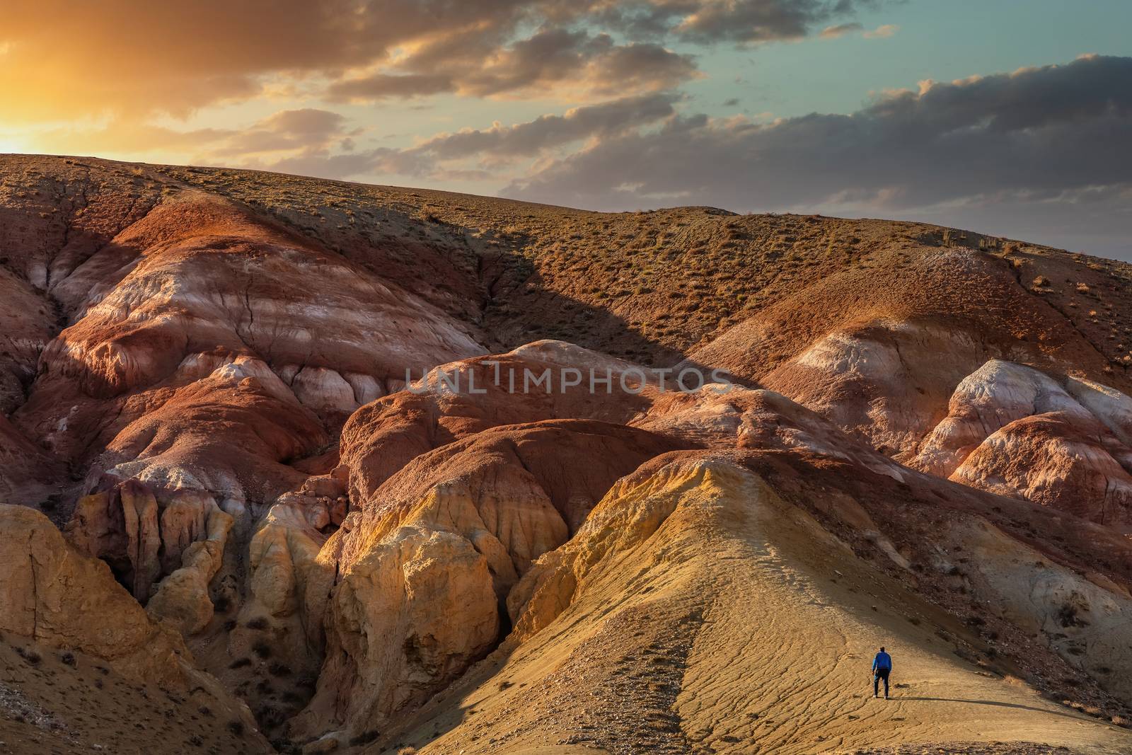 High angle shot of a tourist climbing massive red mountains in Kyzyl-Chin valley, also called Mars valley. Beautiful cloudy sunset sky as a background. Golden hour. Altai, Siberia, Russia.