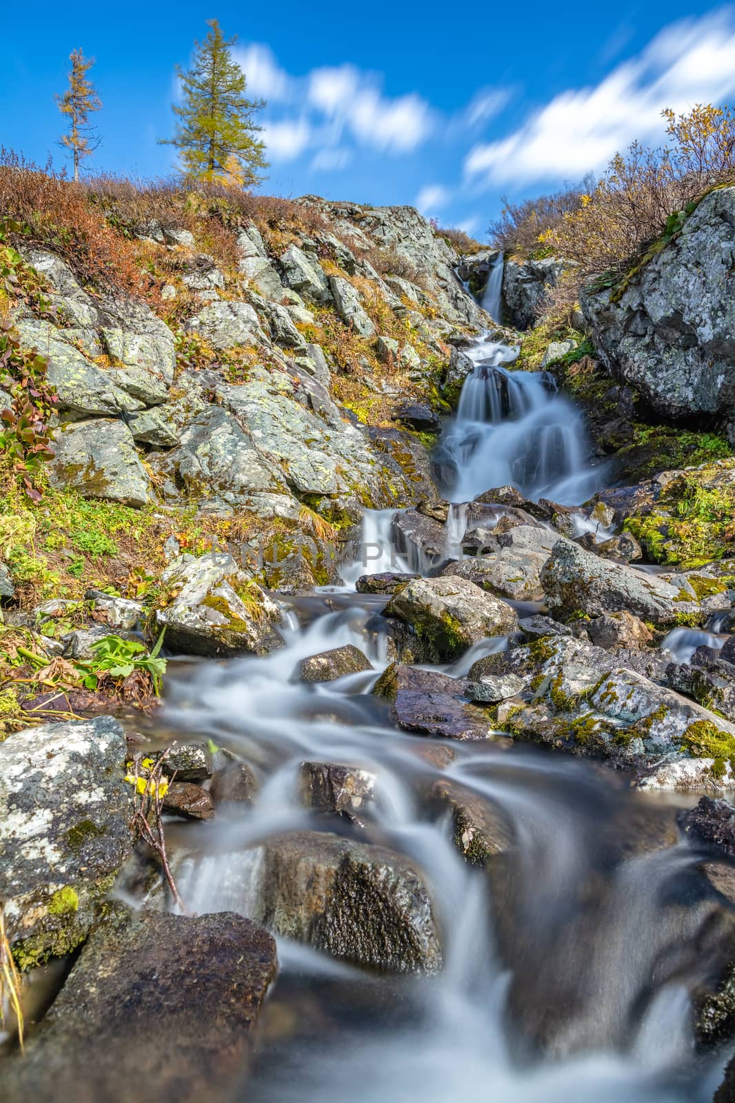 Beautiful low angle shot of waterfall in Altai mountains in Siberia, Russia. Karakolskie lakes. Smooth, silky water and clouds. Blue sky with clouds as a background on top of the hill. Long exposure by DamantisZ