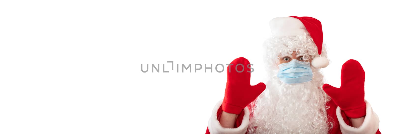 View of a man wearing Santa Claus costume, medical mask, having his both hands up, eyes wide open in warning gesture, isolated on white background. Banner size, copy space. Pandemic holiday concept.