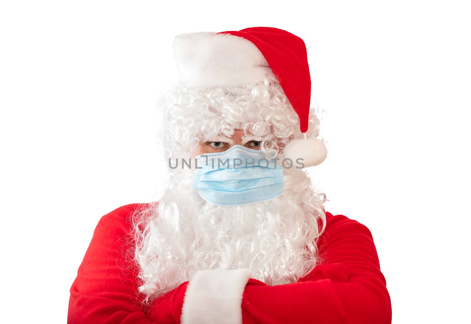 View of a man wearing a Santa Claus costume and medical mask with his arms crossed on his chest, isolated on white background. Man looks very angry. New normal, new reality, pandemic holiday concepts.