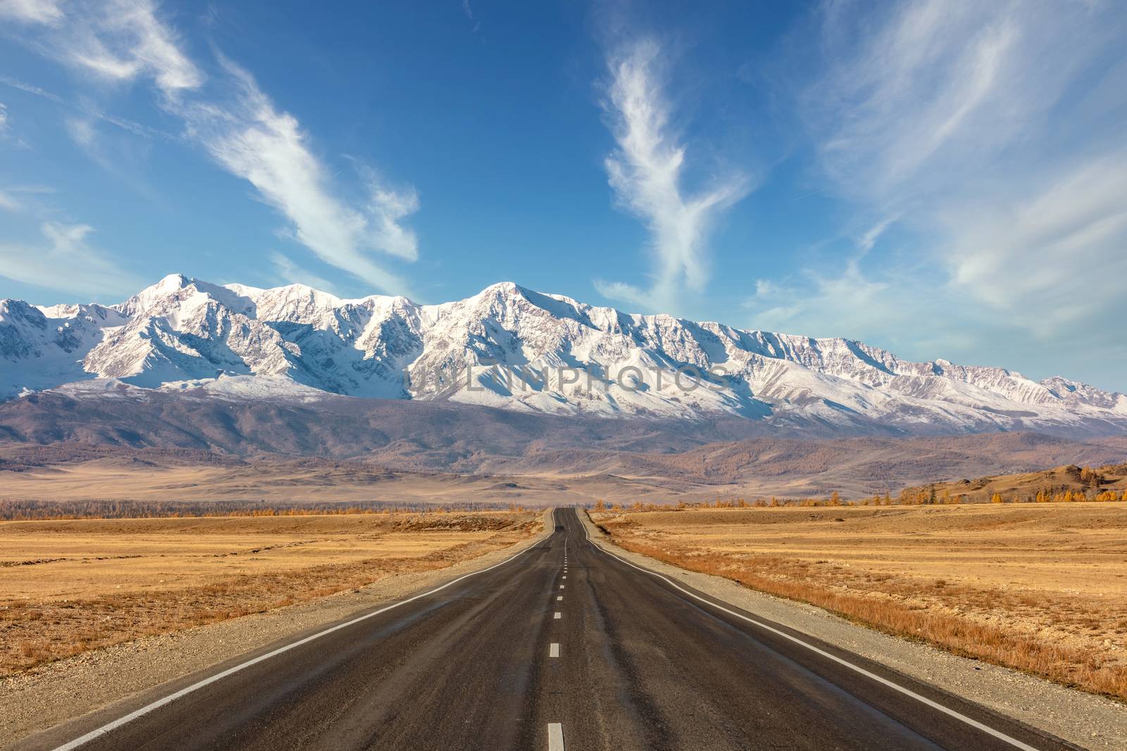 Shot of a straight empty highway leading to the snowy peaks of The Kuray mountain range. Beautiful blue cloudy sky as a background. Altai mountains, Siberia, Russia.