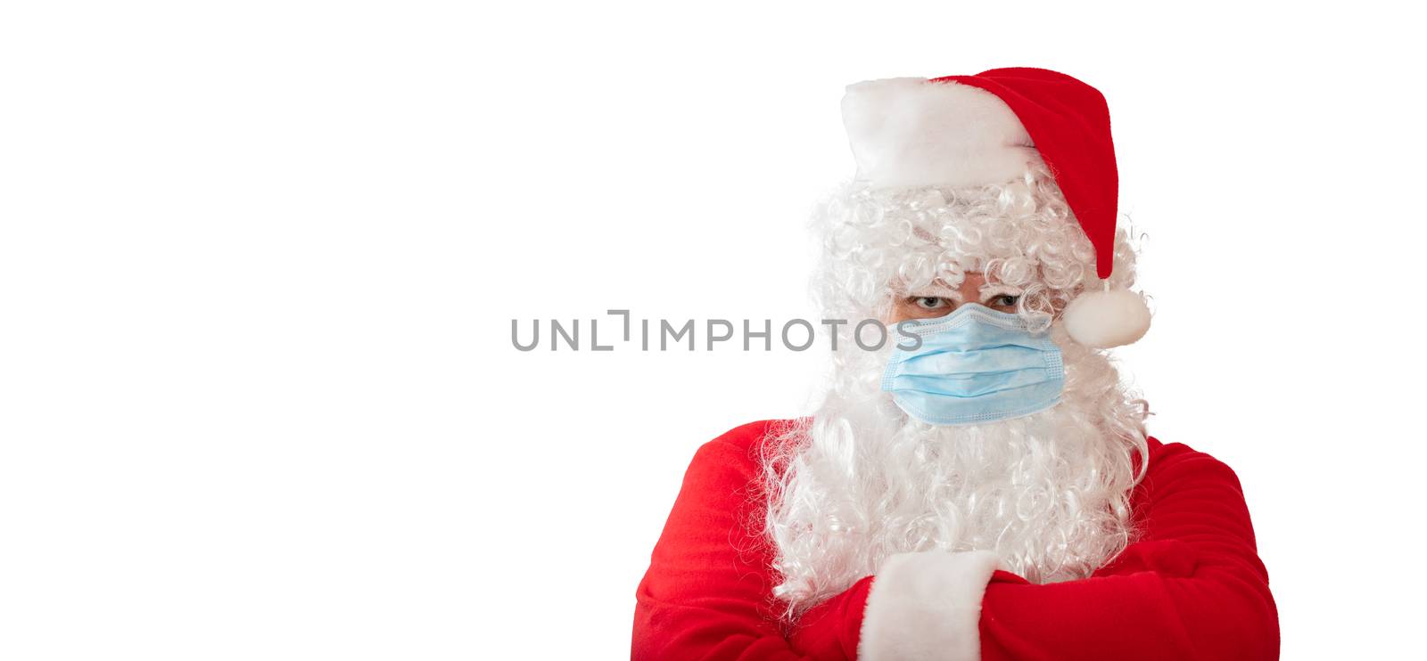 View of a man wearing a Santa Claus costume and medical mask with his arms crossed on his chest, isolated on white background. Banner size, copy space. New normal, pandemic holiday concepts.