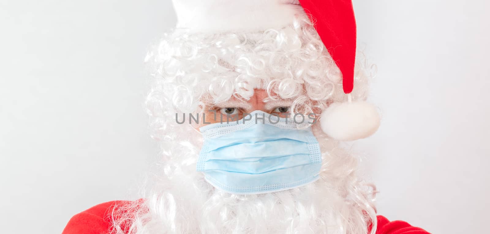 Close-up shot of man wearing a Santa Claus costume and a medical mask on white background. Man looks very angry and stressed, staring straight in the camera. New normal, new reality, holiday concepts by DamantisZ