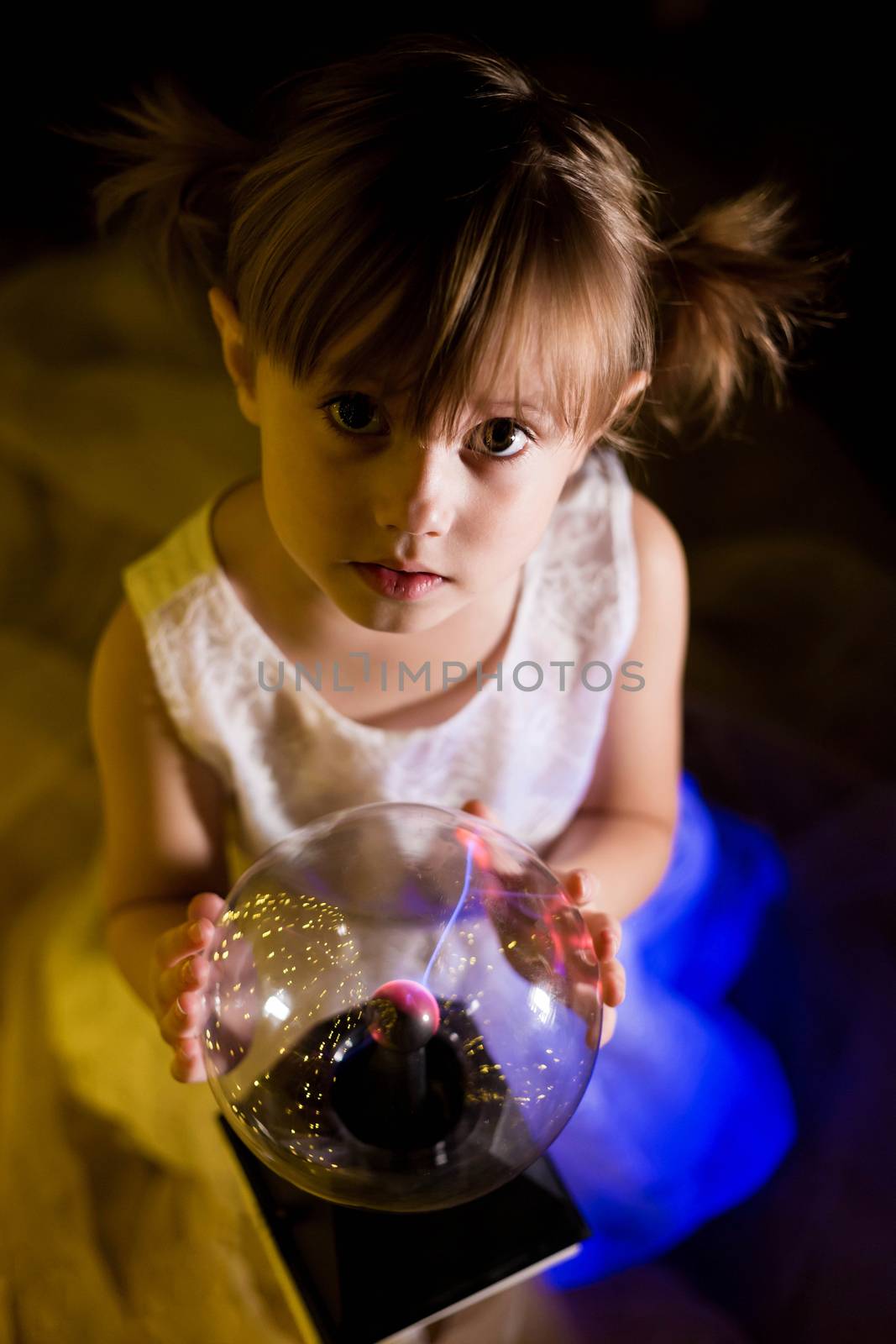 Cute little Child girl holding witch crystal ball with lightning and electricity over dark background with garlands bokeh