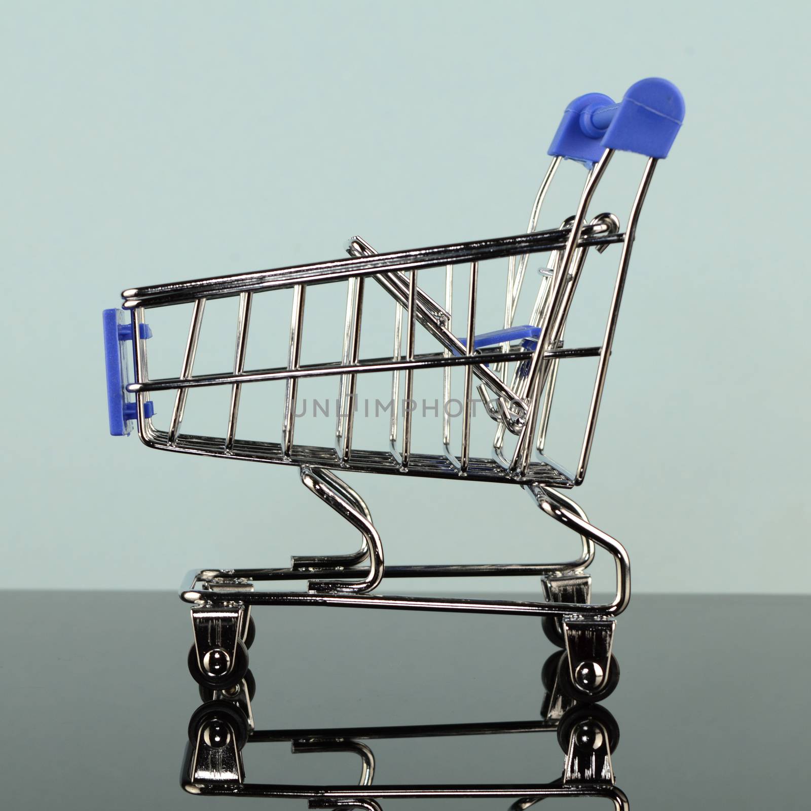 A square format image of a miniature shopping cart over a blue reflective background.