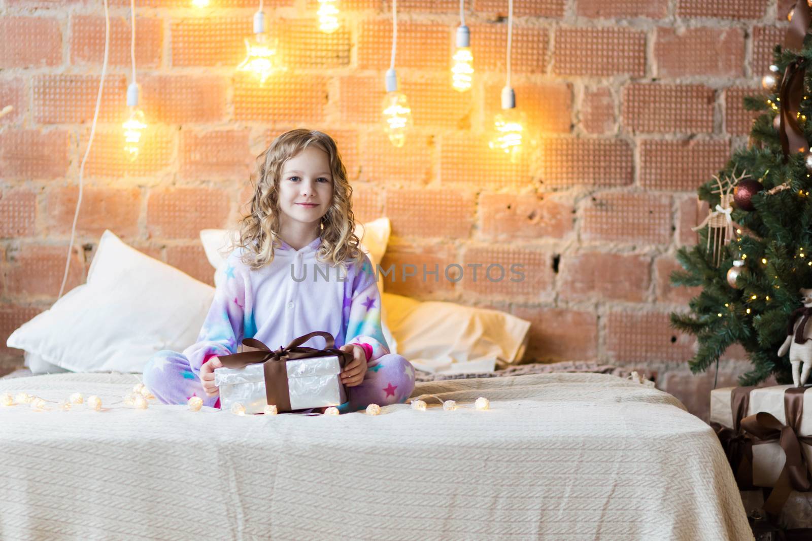 Adorable girl in pajamas sits on the bed and opens a gift against the background of New Year's garlands