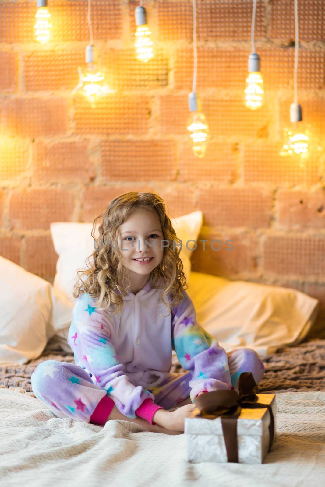 Adorable girl in pajamas sits on the bed and opens a gift against the background of New Year's garlands