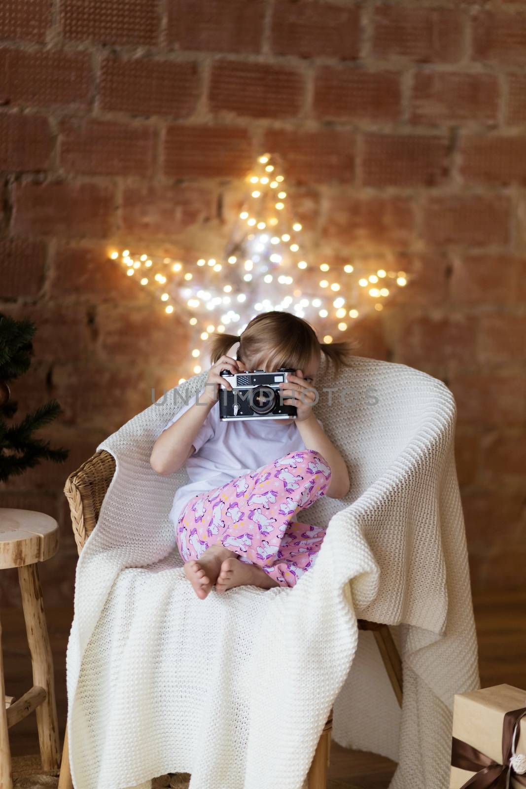 A girl in pajamas sits on an armchair with a camera on the background of garlands