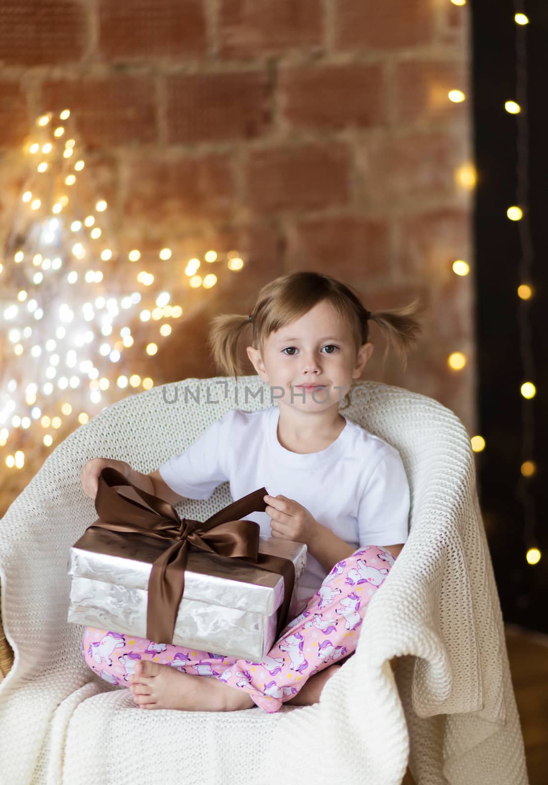 Beautiful child with a christmas gift in pajamas sitting on the chair with gift box looking at the camera