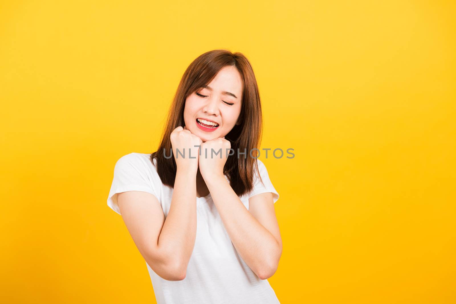 Asian happy portrait beautiful cute young woman teen stand wear t-shirt happy expression fist pressed together under chin and closed eyes isolated, studio shot on yellow background with copy space