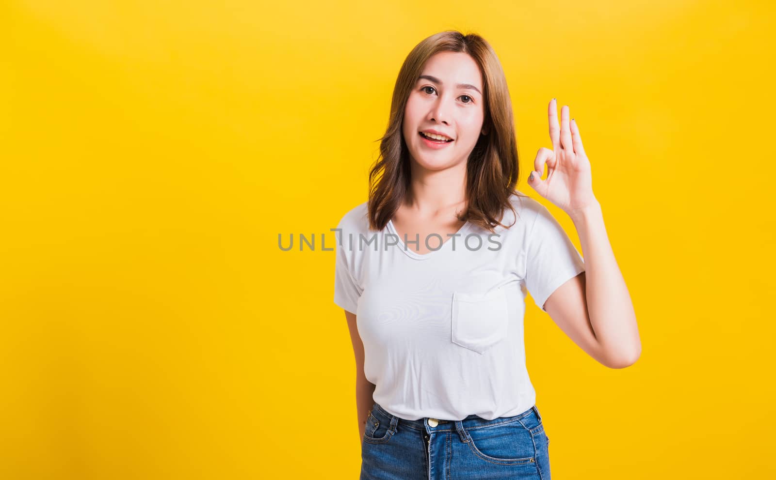 Asian Thai happy portrait beautiful cute young woman standing wear t-shirt showing gesturing ok sign with fingers looking to camera, isolated studio shot on yellow background with copy space