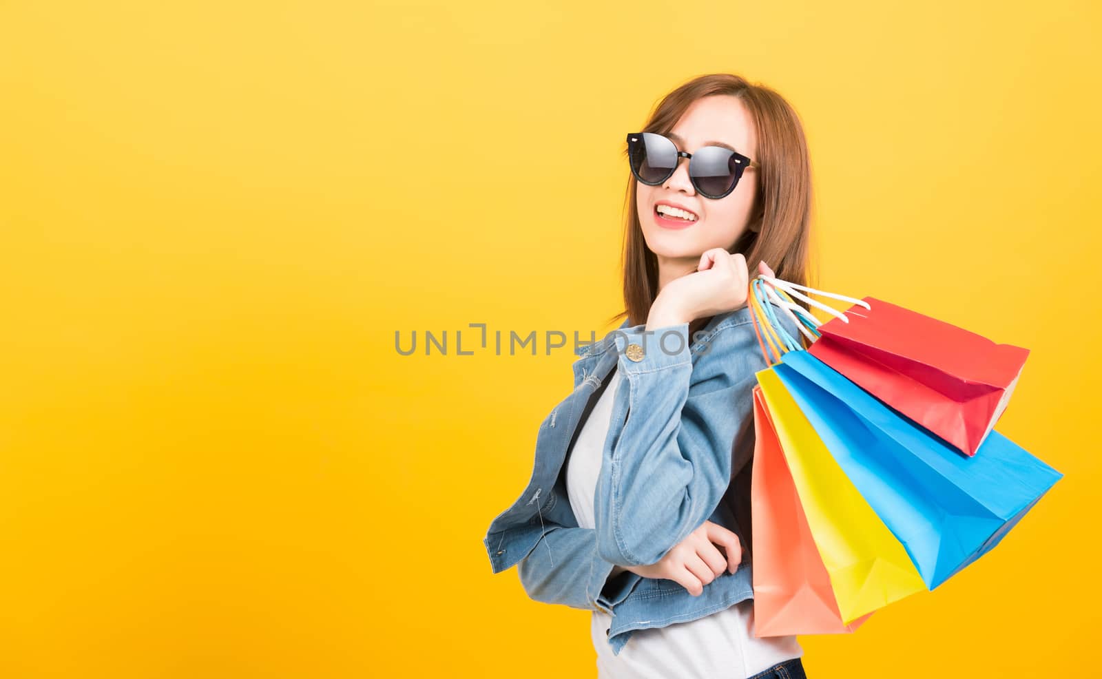 Asian happy portrait beautiful cute young woman teen smiling standing with sunglasses excited holding shopping bags multi color looking up isolated, studio shot yellow background with copy space