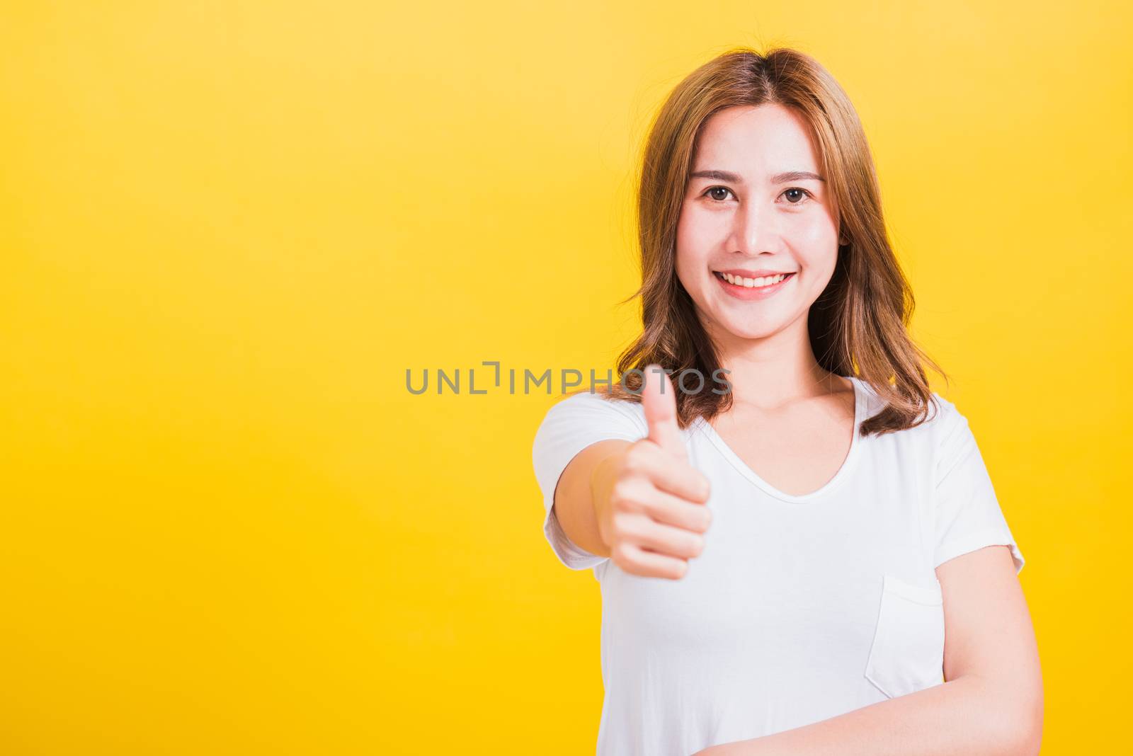 Portrait Asian Thai beautiful happy young woman smiling wear white t-shirt standing successful woman giving one thumbs up gesture sign in studio shot, isolated on yellow background with copy space