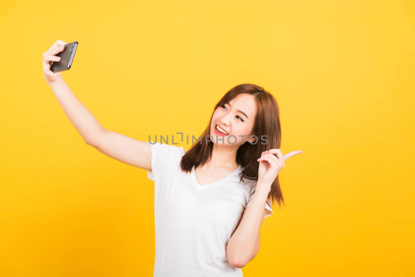 Asian happy portrait beautiful cute young woman teen smiling standing wear t-shirt mak selfie photo, video call on smartphone, point finger back isolated, studio shot yellow background with copy space