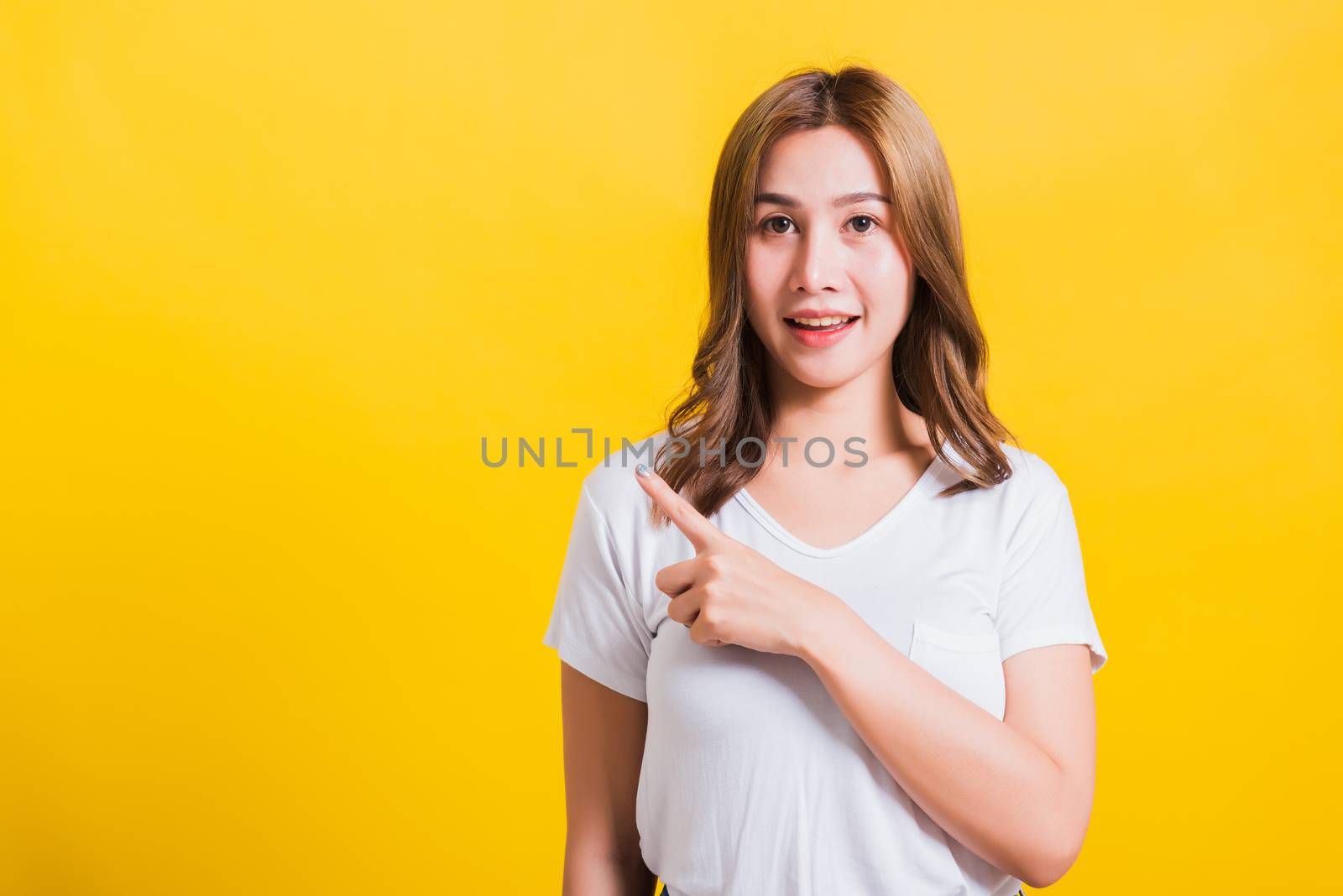 Asian Thai happy portrait beautiful cute young woman standing wear white t-shirt pointing finger away side looking to away side, studio shot isolated on yellow background with copy space