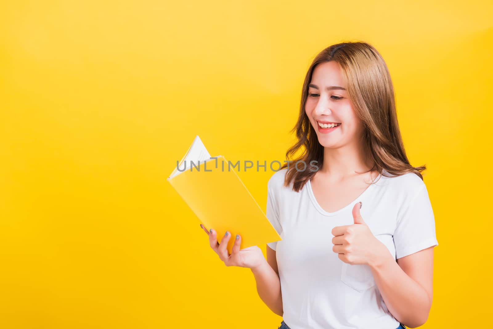 Portrait Asian Thai beautiful happy young lifestyle woman stands holding yellow book or diary she show thumb up finger and looking to book, studio shot isolated on yellow background, with copy space