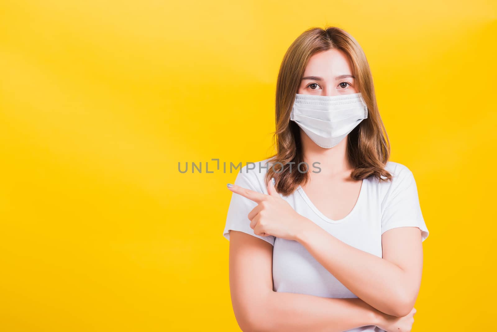 Portrait Asian beautiful young woman wearing face mask protects filter dust pm2.5 anti-pollution, anti-smog, air pollution and COVID virus her pointing out side on yellow background, with copy space