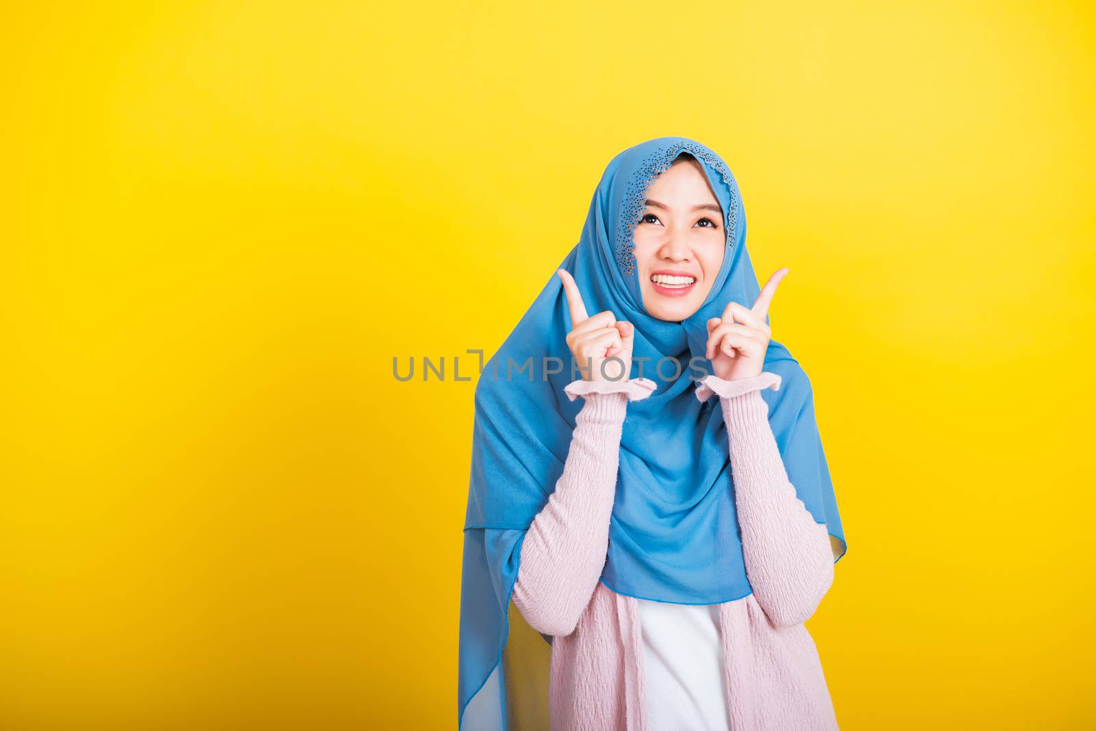 Asian Muslim Arab, Portrait of happy beautiful young woman Islam religious wear veil hijab funny smile she positive expression pointing with finger up to space isolated yellow background