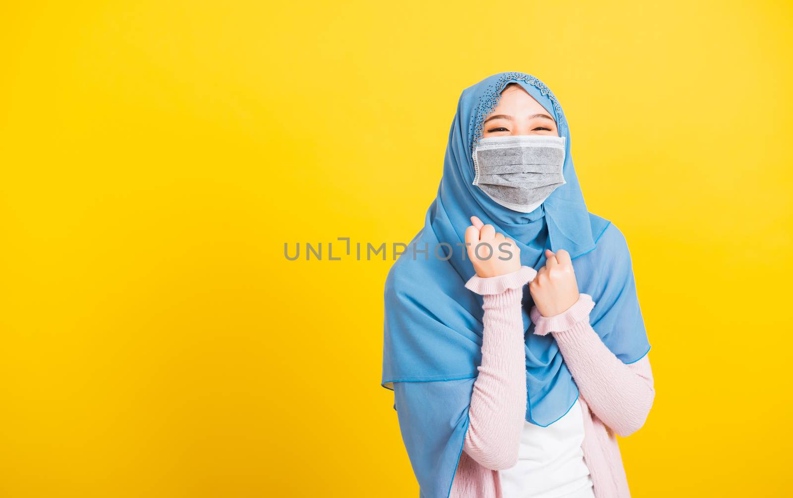 Muslim Arab, Happy beautiful young woman wear veil hijab wear protective face mask quarantines coronavirus raises hands glad excited cheerful after recovering from illness, isolated yellow background