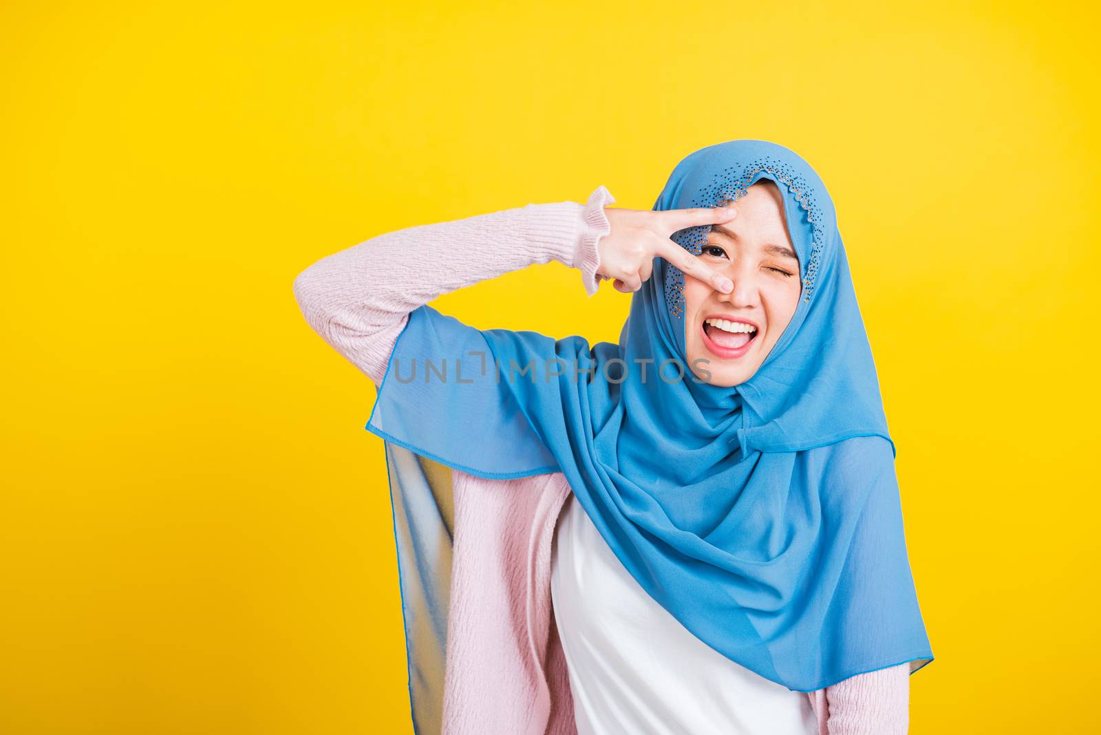 Asian Muslim Arab, Portrait of happy beautiful young woman Islam religious wear veil hijab smile she showing gesture hand two finger V-sign symbol near eyes isolated yellow background