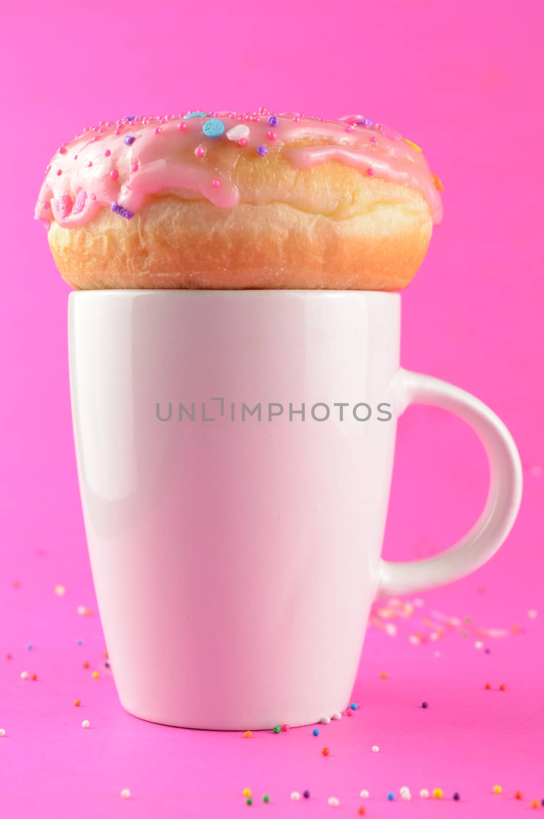 A vertical composition of a coffee cup with a frosted donut resting ontop for the morning breaktime.