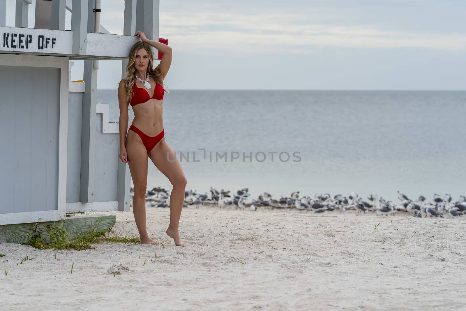 A beautiful blonde bikini model enjoys the weather outdoors on the beach while posing near a lifeguard station