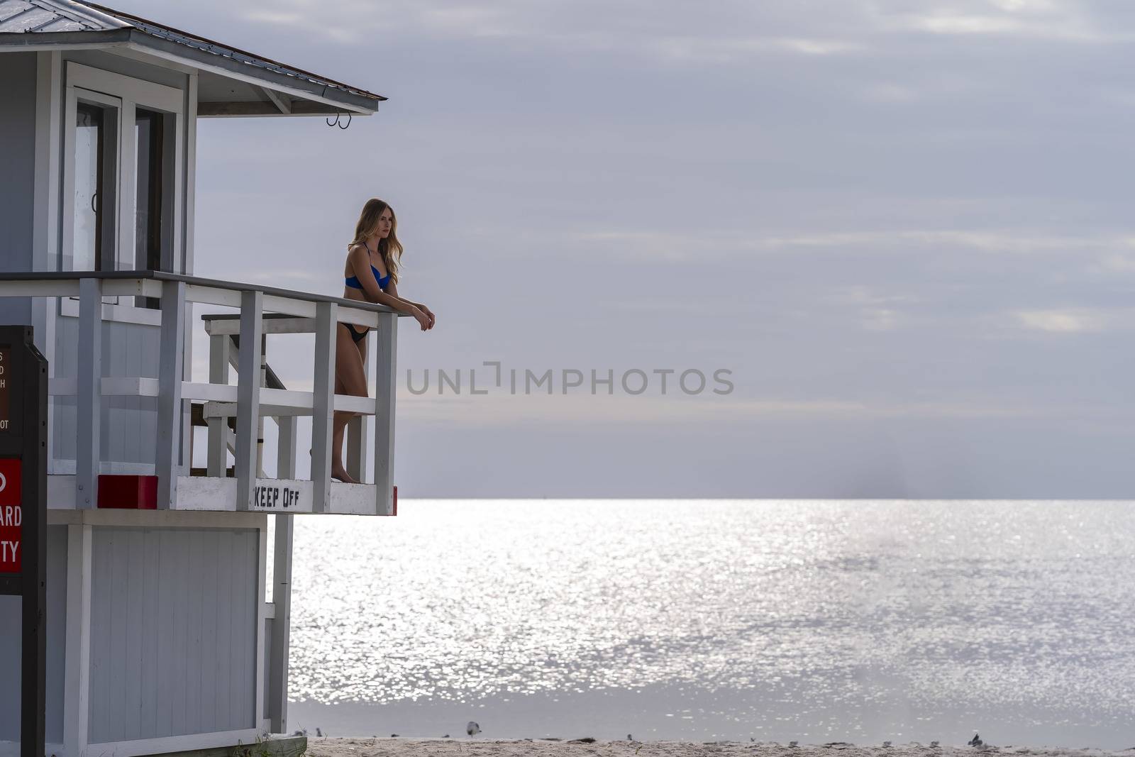 A beautiful blonde bikini model enjoys the weather outdoors on the beach while posing near a lifeguard station