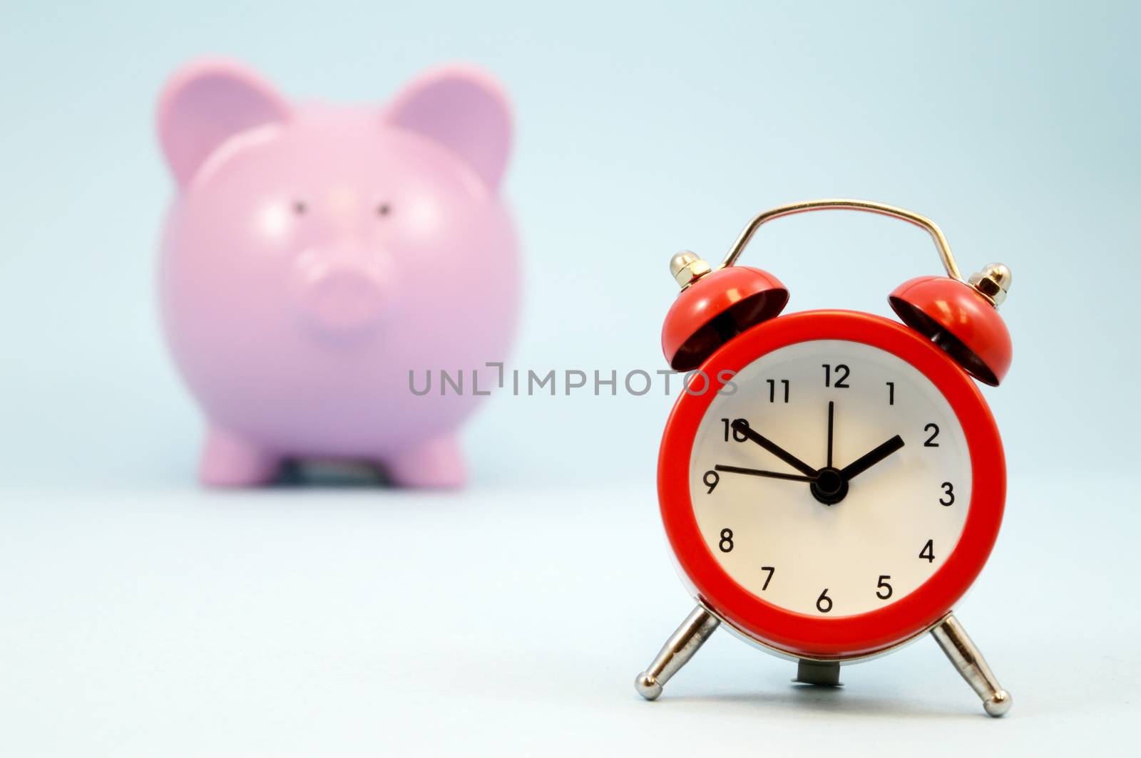 A red alarm clock focused in the forground with a blurred pink piggy coin bank in the background all over blue.