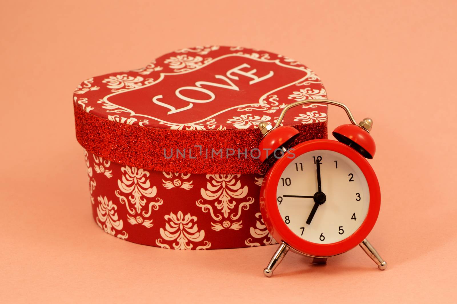 A red heart shaped gift box and an alarm clock over a pink background remind a romantic couple of important times.