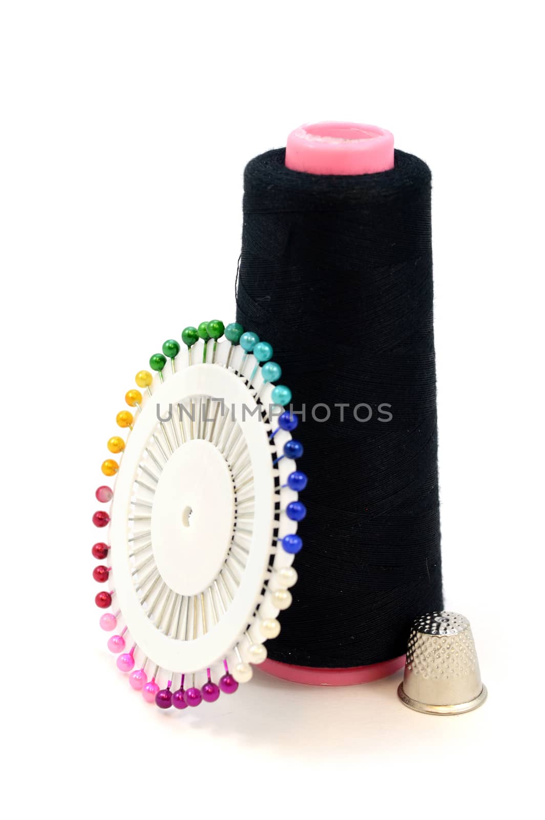 An isolated still-life of sewing objects over a white background including a spool of black thread with multicolor pins and a metal thimble.