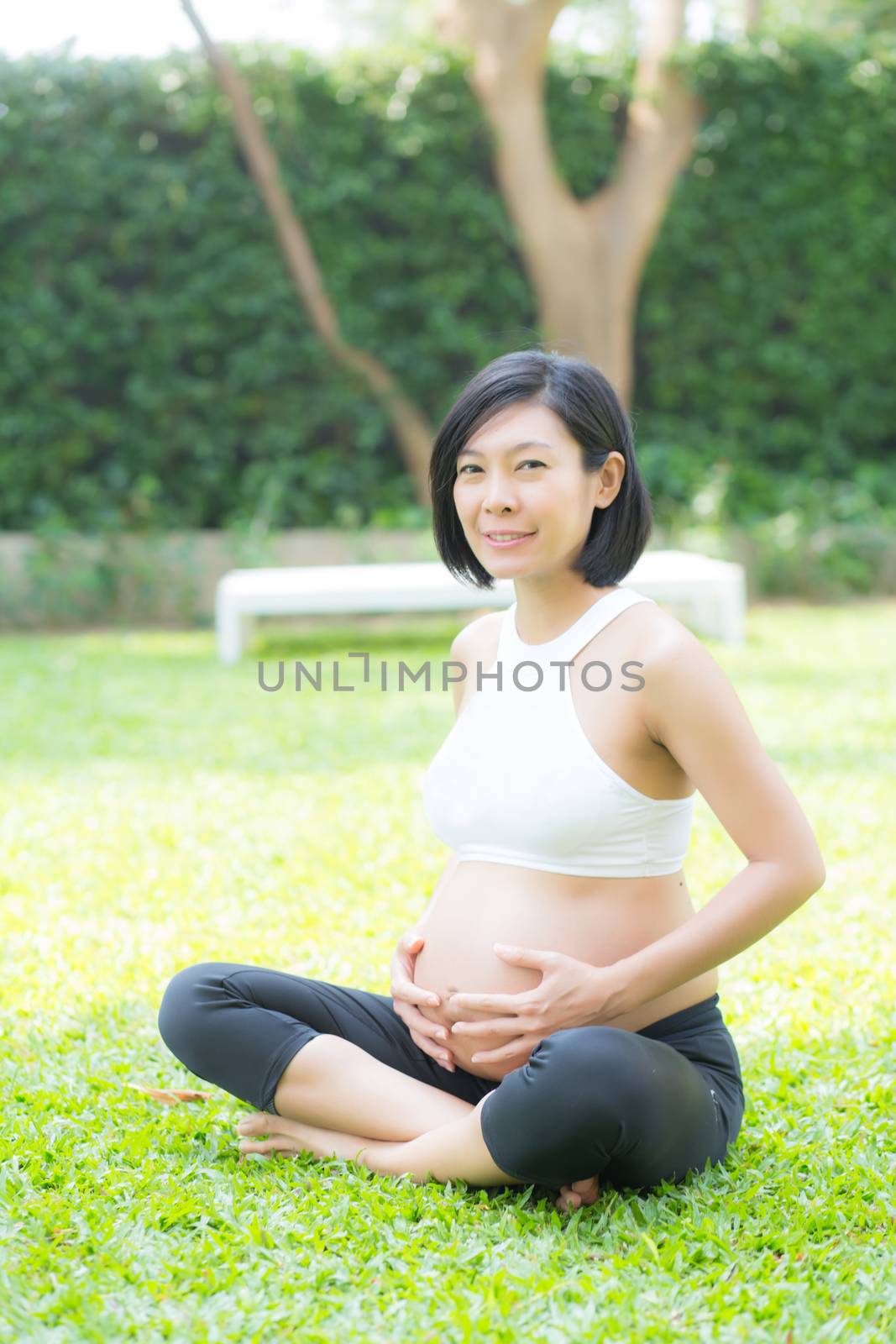 Beautiful pregnant asian young woman relax in the park, girl with belly sitting with crossed leg on grass and yoga exercise for wellness, copy space.