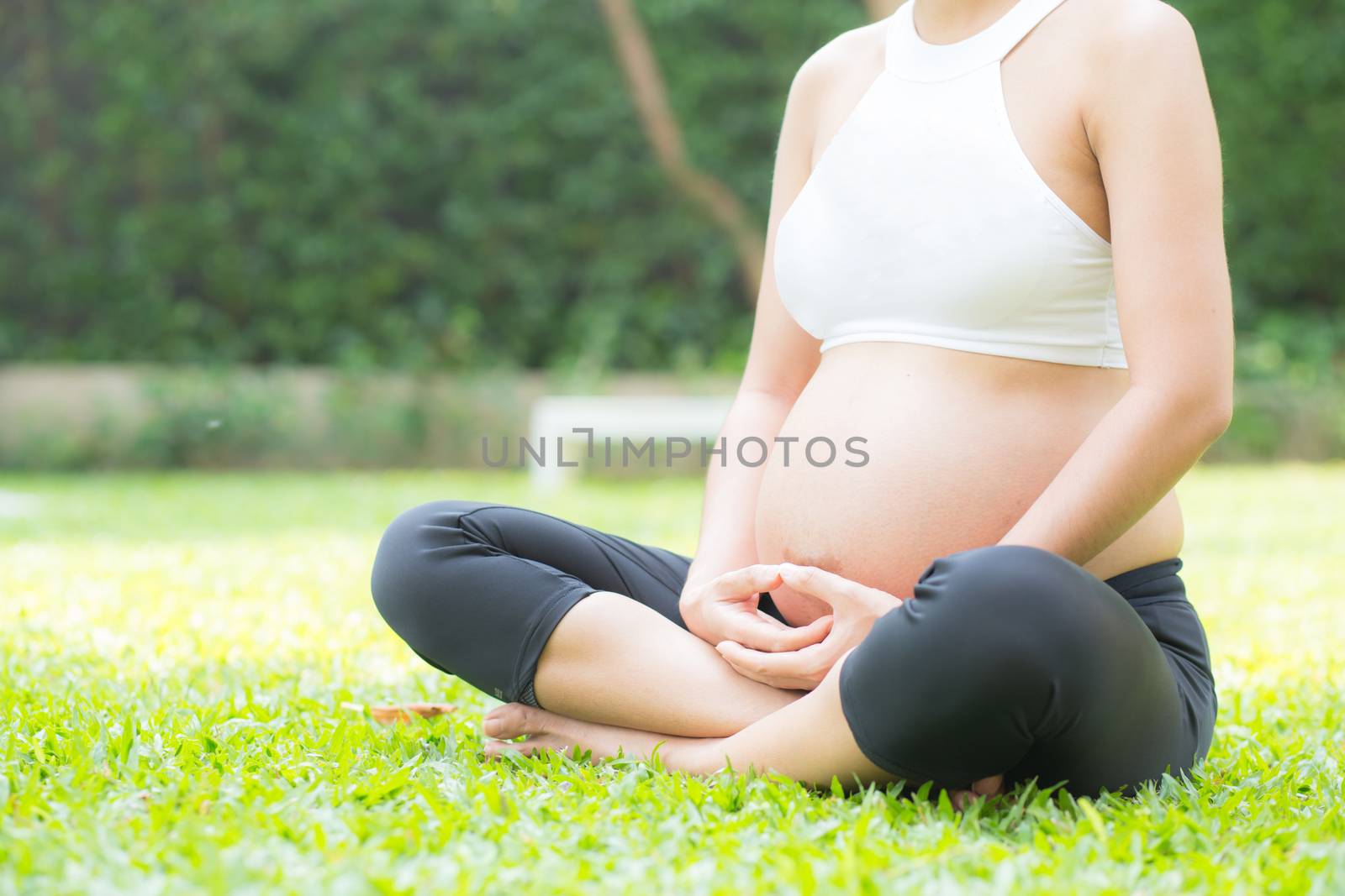 Beautiful pregnant asian young woman relax in the park, girl with belly sitting with crossed leg on grass and yoga exercise for wellness, copy space.