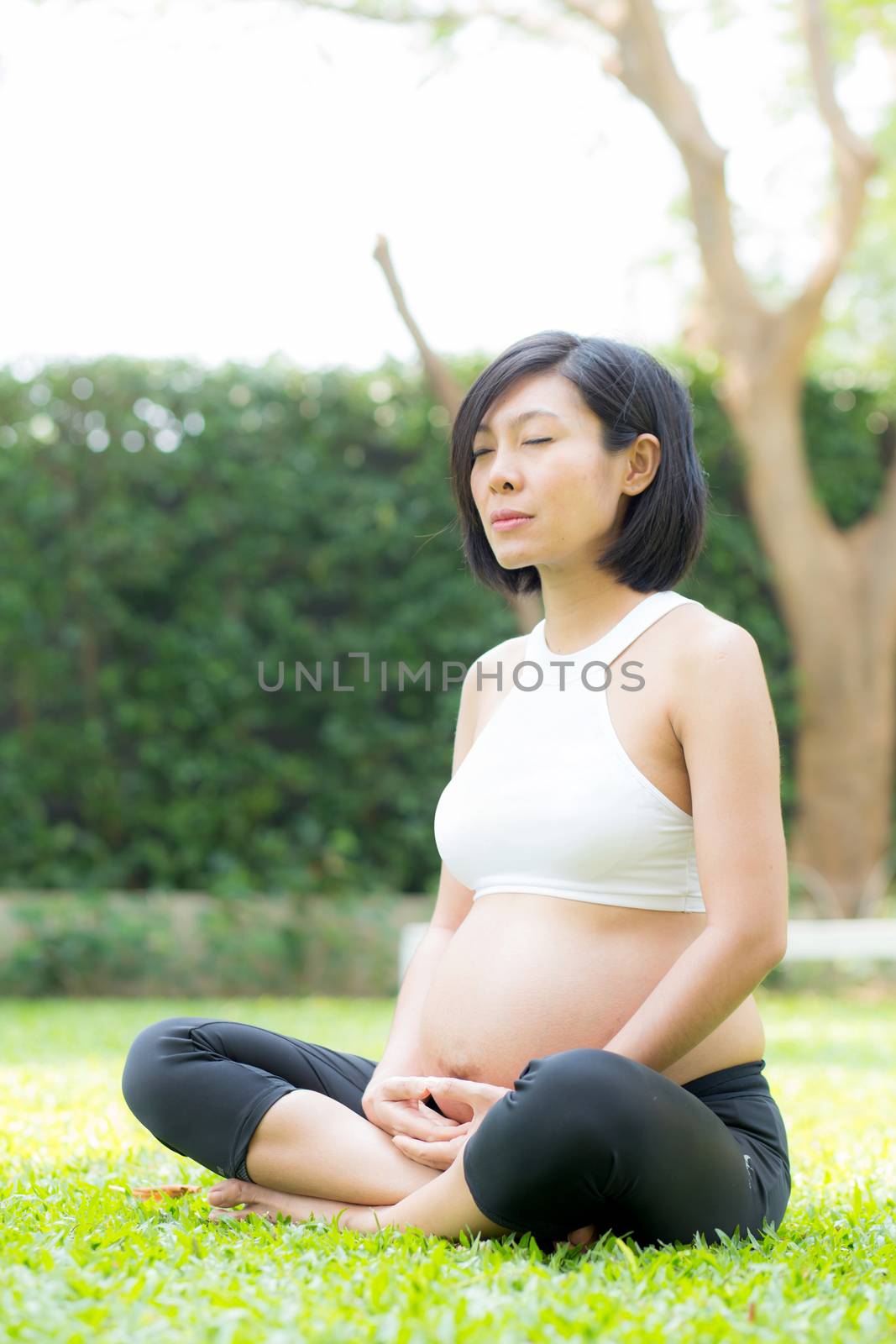 Beautiful pregnant asian young woman relax in the park, girl with belly sitting with crossed leg on grass and yoga exercise for wellness, copy space.